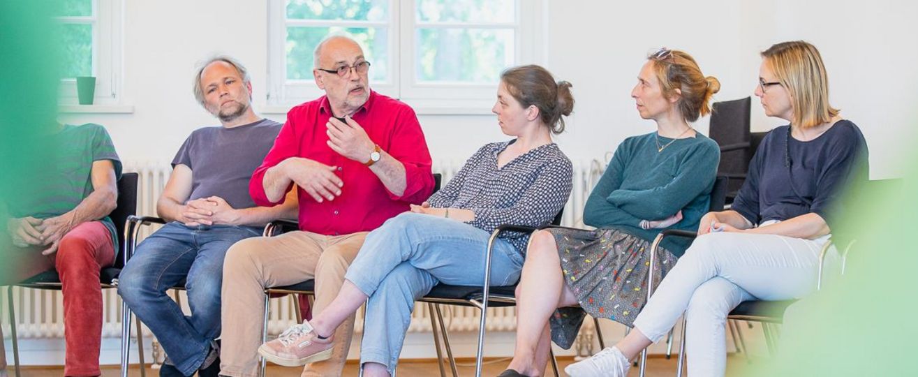 6 Menschen sitzen als Teil einen große Stuhlkreises und sprechen miteinander