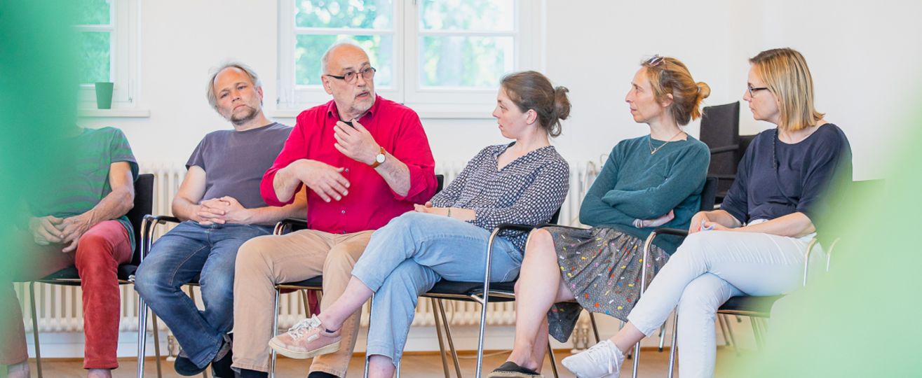 6 Menschen sitzen als Teil einen große Stuhlkreises und sprechen miteinander