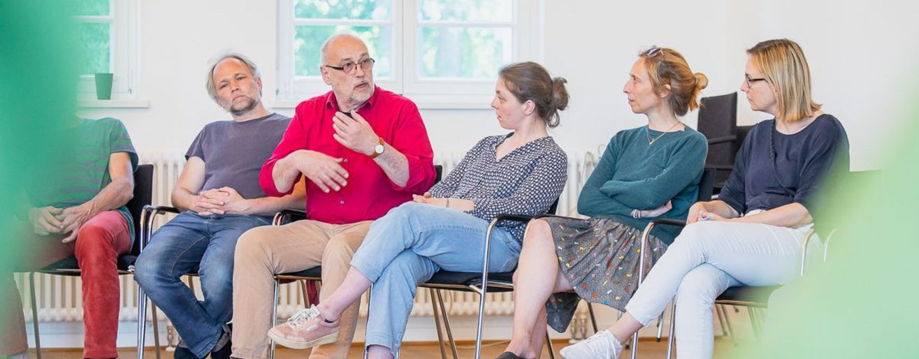 6 Menschen sitzen als Teil einen große Stuhlkreises und sprechen miteinander