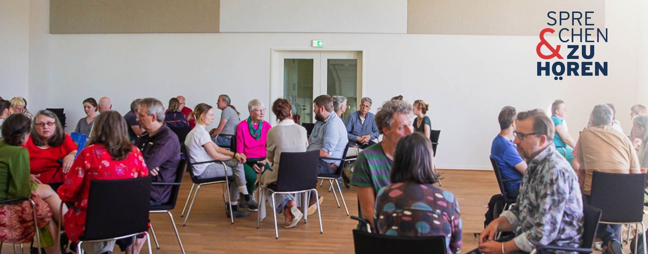 Foto einer Sprechen-und-Zuhören-Veranstaltung mit Menschen auf Stühlen in Gruppen sitzend
