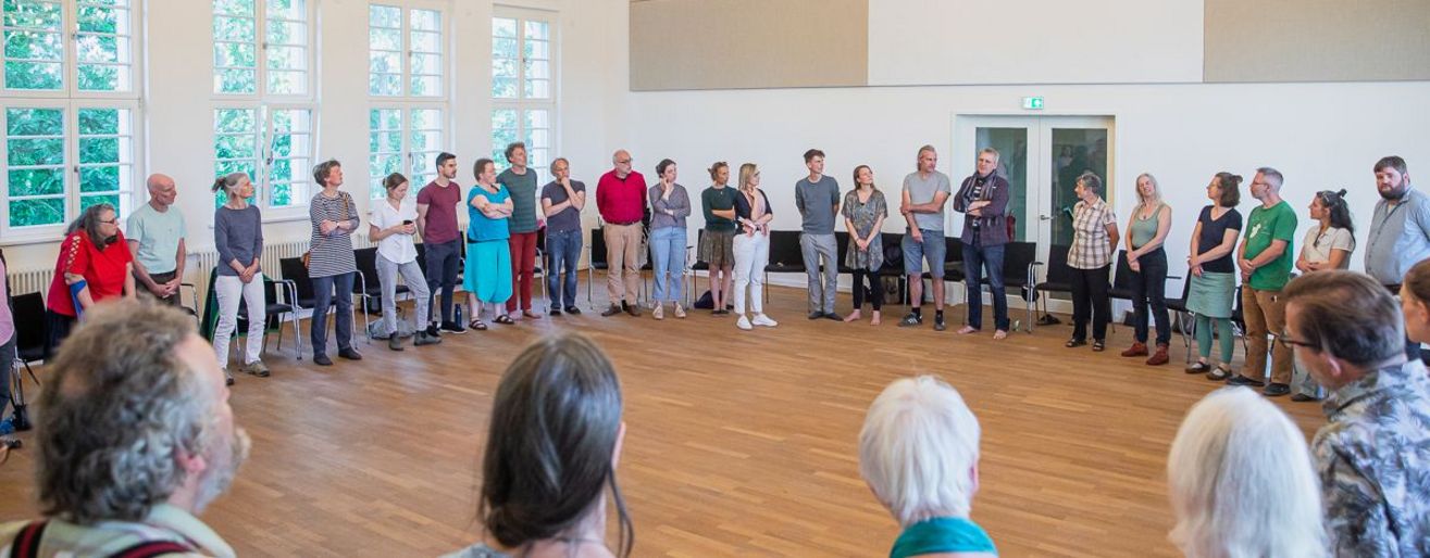 Circa 50 Menschen sitzen in einem großen Stuhlkreis in einer Halle.