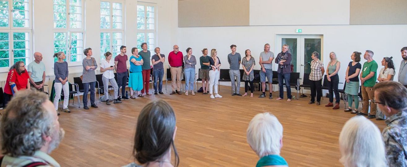 Circa 50 Menschen sitzen in einem großen Stuhlkreis in einer Halle.