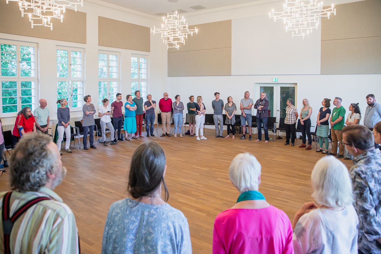 Ein Saal mit 40 Menschen die im Kreis stehen.